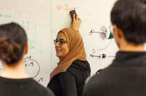 woman writing equations on white board while two students watch