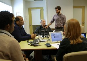 Group sitting around table watching man hold device against chest of another man who is sitting