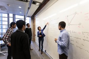 Female professor writing on whiteboard with students surrounding her