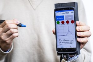 Closeup of person’s hands holding rubber-tipped ear canal probe and smartphone attached to 3D-printed casing. Smartphone screen shows tympanometry software interface displaying current action as “Measure,” with “Stop” grayed out, and a peaking line graph with x axis scale from -400 to 200 and y axis scale of -0.5 to 1.5. Above the graph is a row of color-coded circles with indicators for “connected” (green), and “measuring,” “seal, ”and “reset” (red). Below the graph is text: “Peak admittance: 0.8 ml, Ear canal volume: 0.4 ml,” Peak pressure: 0 daPa.”