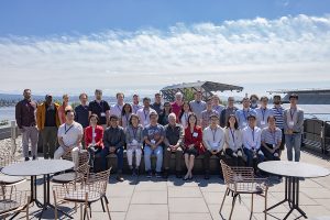 Large group of people sitting, posing for a photo on the rooftop deck of the Bill & Melinda Gates Center for Computer Science & Engineering, with Husky Stadium in the background