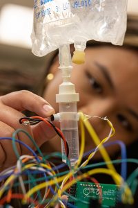 A woman adjusts the drip rate of an IV bag attached to wires and circuitry