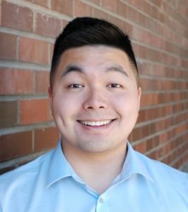 Headshot of UW ECE alum Frank Liu in a light blue collared button up standing in front of a red brick wall. 