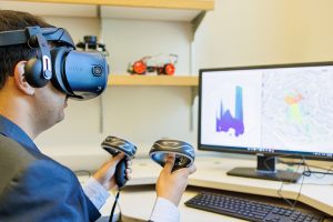 professor Akshay Gadre wearing an AR/VR headset, holding controllers, while sitting in front of a computer screen showing a blueprint map of the UW campus