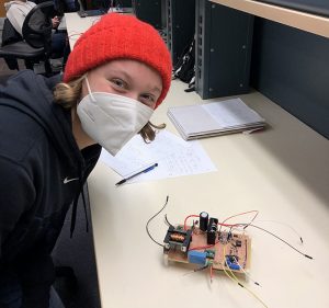 Margot Adam, wearing a ski hat and COVID facial mask, sitting next to a circuit board on a desk