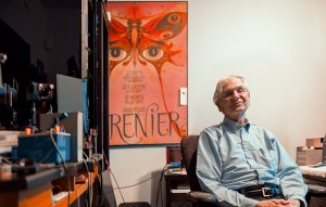 UW ECE Affiliate Professor Gary Bernard sitting in his home laboratory next to a poster that shows an illustration of a butterfly