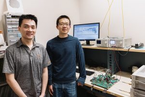 Two men standing in front of a desk with a complex circuit board on it.