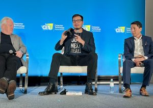 Three men sit in chairs on a stage in front of a screen at the 2024 CES. The man in the center, Alvin Graylin, holds a virtual reality headset.