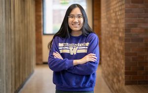 UW ECE undergraduate Mary Bun stands in a hallway, smiling, with arms folded across her chest. She is wearing glasses and a purple sweatshirt.
