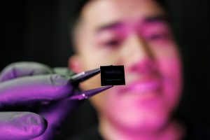Bingzhao Li holding a microchip with tweezers. He is smiling and the chip is close to the camera.