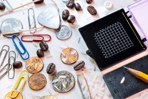 Coins, paperclips, coffee beans, a ruler, pencil, and grains of rice jumbled together on a white surface, next to a black and white grid containing a tiny microchip