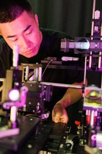 Bingzhao Li in the lab, looking into a microscope. He is surrounded by lab equipment.
