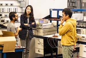 UW ECE Assistant Professor Jungwon Choi standing in between two graduate students with her arms crossed, in conversation. The room is full of boxes, wires, and electronic equipment.