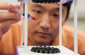 A needle adds a drop beneath four other drops suspended in middair The drops are within an acoustic levitator and behind the scene is the blurred face of Jared Nakahara, who is the one holding the needle.