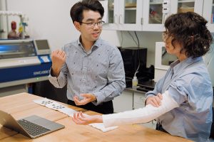 Sen Zhang standing at a table with Yiyue Luo. Yiyue is wearing a MultiSensKnit prototype on her arm. On the table is another MultiSensKnit prototype.