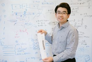 Sen Zhang holds a white knitted sleeve with a gray strip running down the middle. Behind him is a white board containing all sorts of calculations.