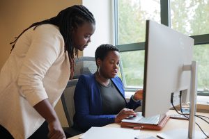 UW ECE Assistant Professor June Lukuyu standing next to Eliane Nirere, who is sitting and working on a computer on a desk by a window.
