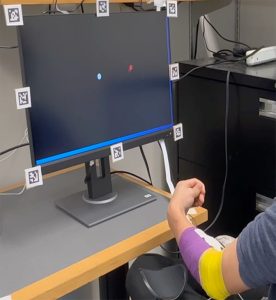 A person's arm, wrapped with purple and yellow tape, gestures in front of a black monitor displaying a red and blue dot.