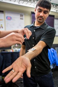 Shwetak Patel holding out his arm, while a pair of hands place a device on his forearm.