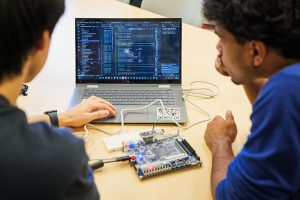 Two students sitting in front of a laptop, which is connected by cables to a microchip.