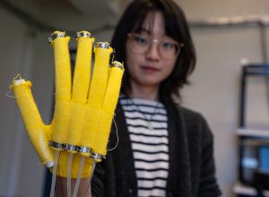 UW ECE Assistant Professor Yiyue Luo wearing a yellow, knitted glove that is embedded with pneumatic actuators. the glove is big and in the foreground of the photo.