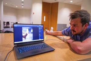 Jayce Gaddis crouched down next to his laptop, which is sitting on a table. Onscreen is a video capture of his dog.